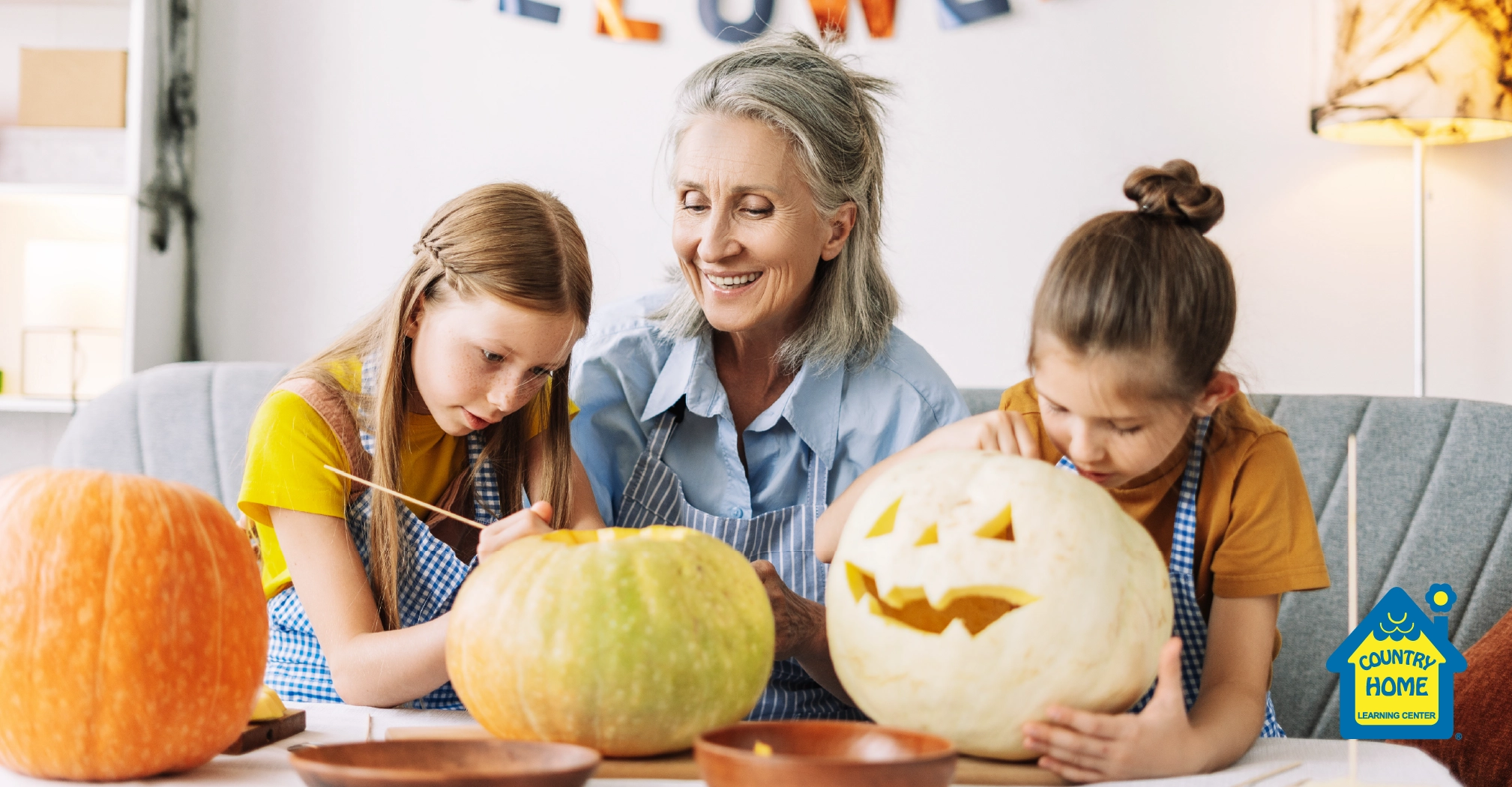 An older woman and two young girls sit at a table carving pumpkins, sharing fun jack-o-lantern ideas. The girls concentrate on their creations while the woman smiles.