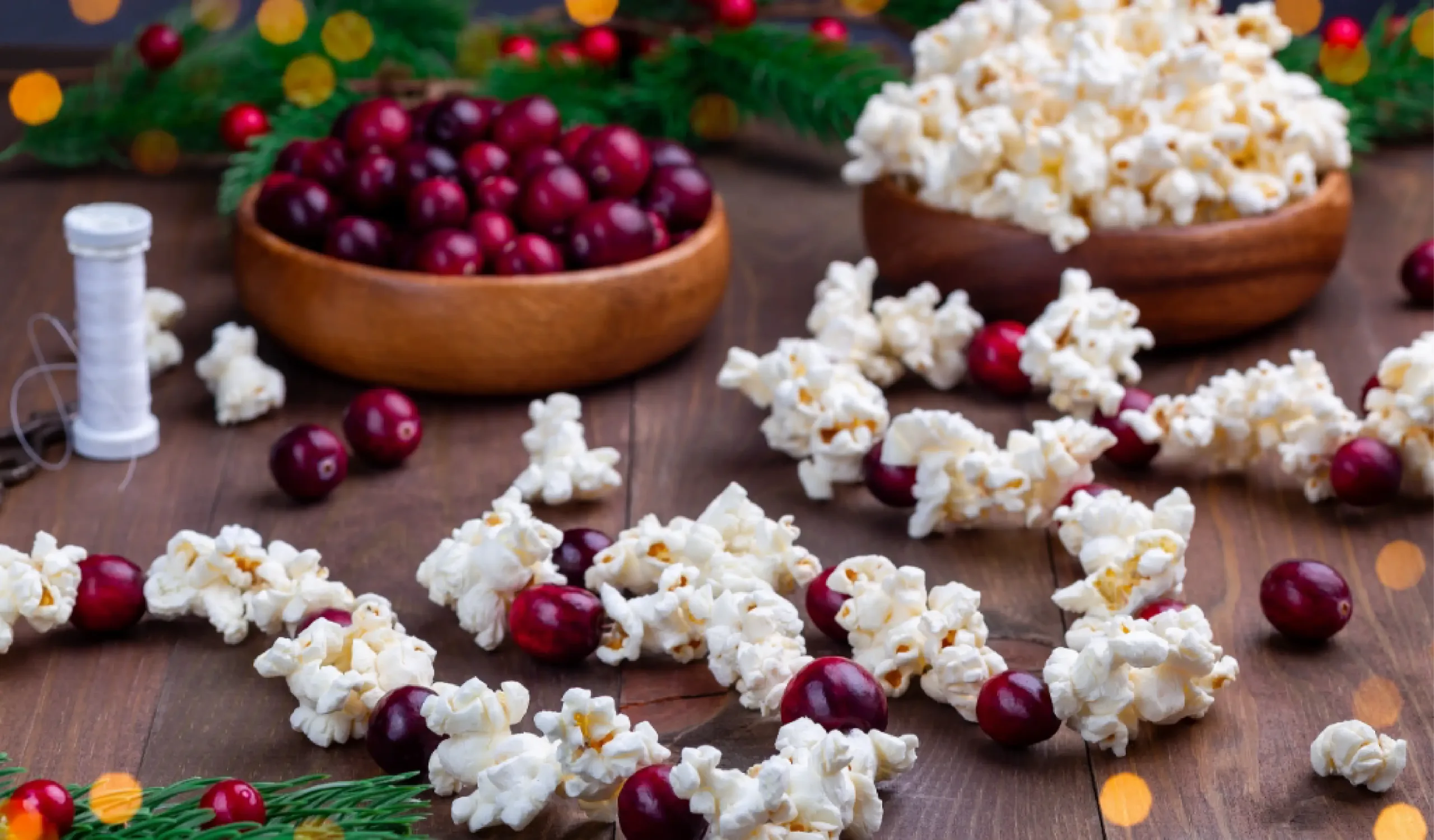 A wooden table with bowls of cranberries and popcorn, a string of cranberries and popcorn—perfect for sharing fun Christmas facts for kids—plus pine branches and a spool of thread in the background.