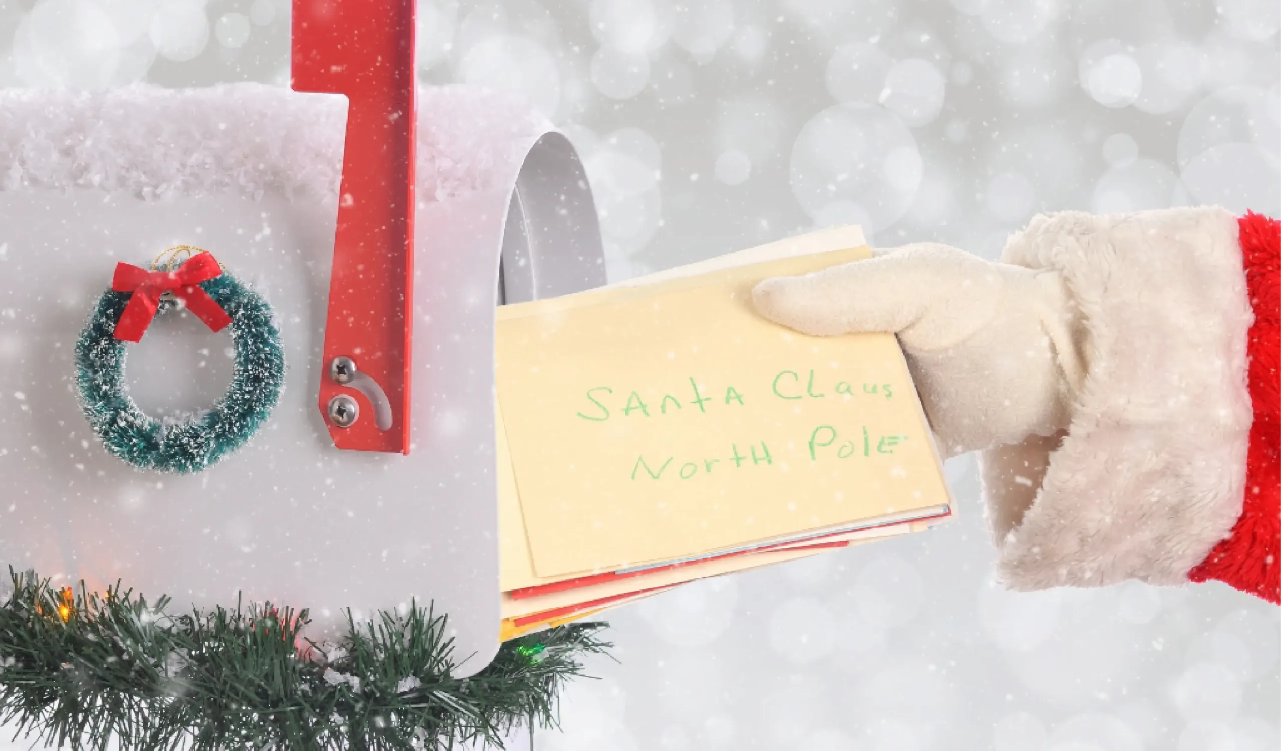 A gloved hand places an envelope labeled "Santa Claus, North Pole" into a white mailbox decorated with a small wreath and greenery in falling snow.