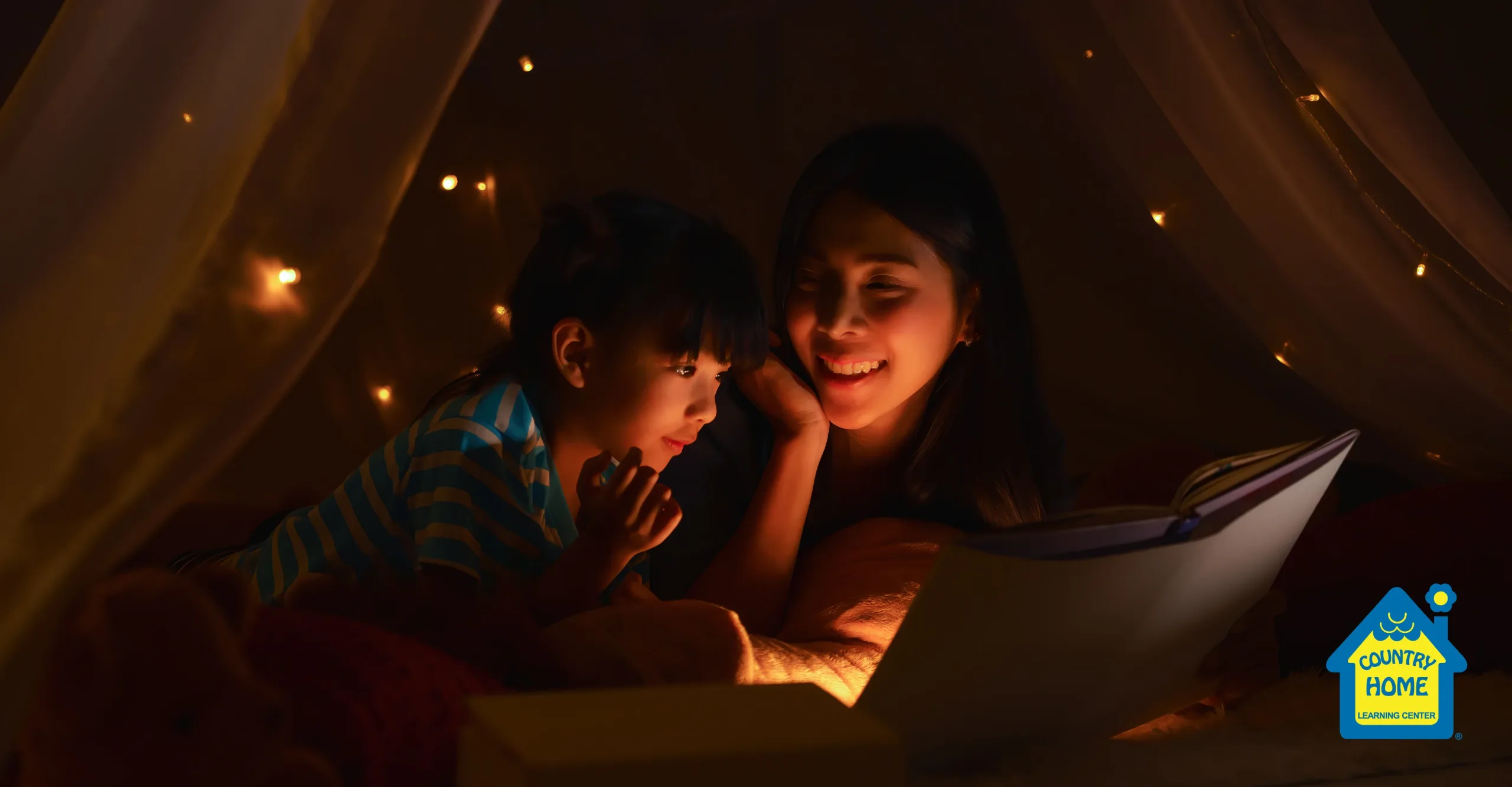 asian mother and daughter spend quality time together inside on a rainy day while reading a book in a tent under twinkly lights