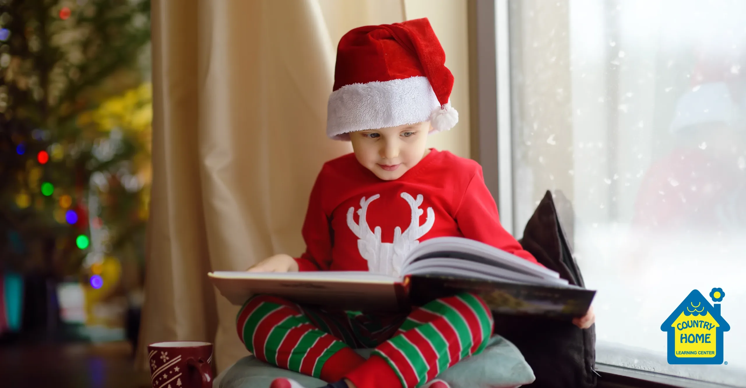 a young boy wearing christmas pajamas and santa hand reads a holiday book next to the christmas tree as it snows outside.