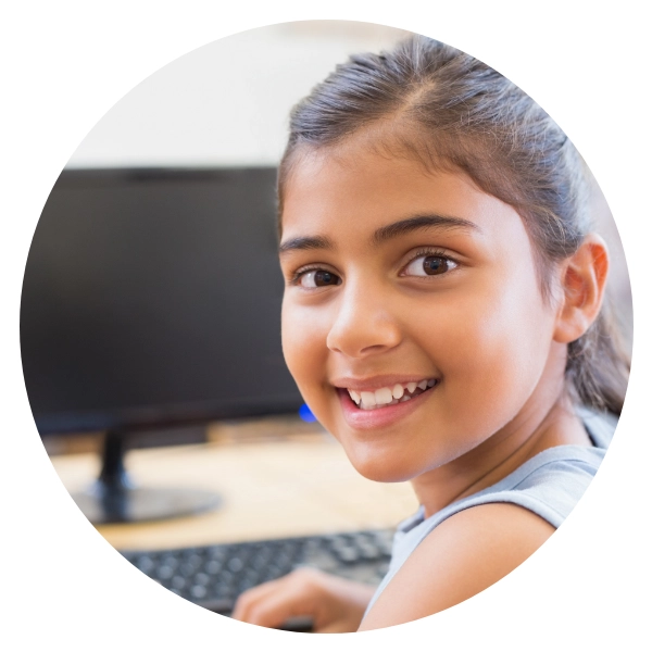 A young girl sits at a computer and smiles at the camera.