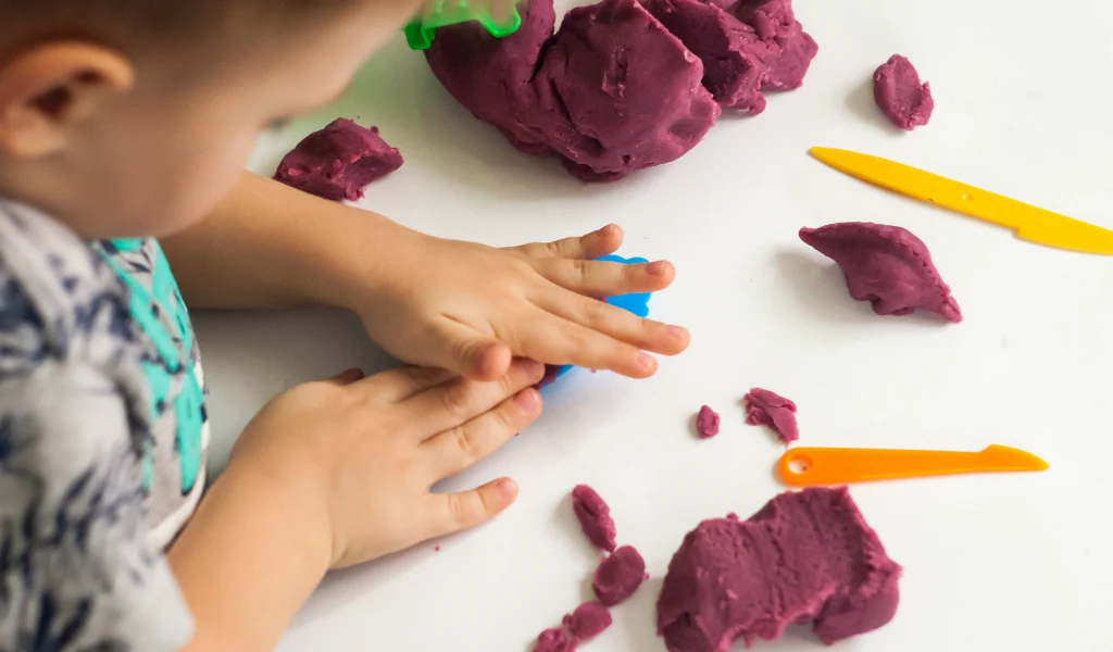 A child molds purple playdough with hands on a table scattered with purple playdough and yellow plastic tools.