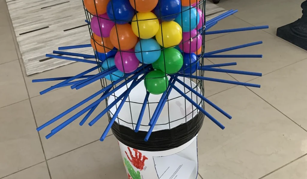 A homemade KerPlunk game with colored plastic balls held in a wire basket by blue sticks above a white bucket.