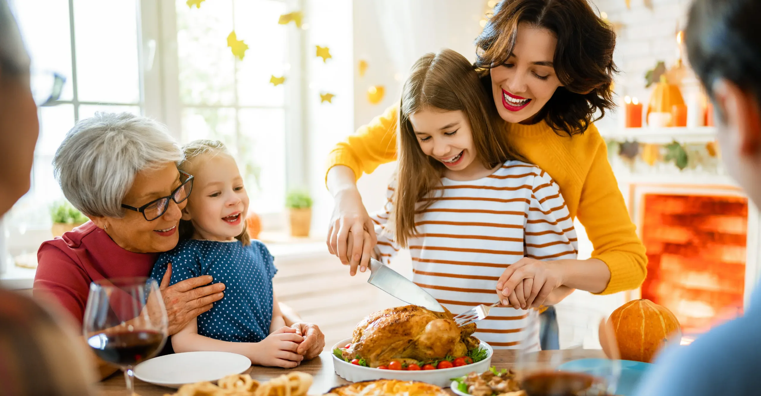 A joyful multigenerational family gathered around a table for a Thanksgiving meal. A smiling mother helps her daughter carve a roasted turkey while a grandmother embraces two young girls. The room is warmly decorated with pumpkins and fall leaves, creating a cozy, festive atmosphere.