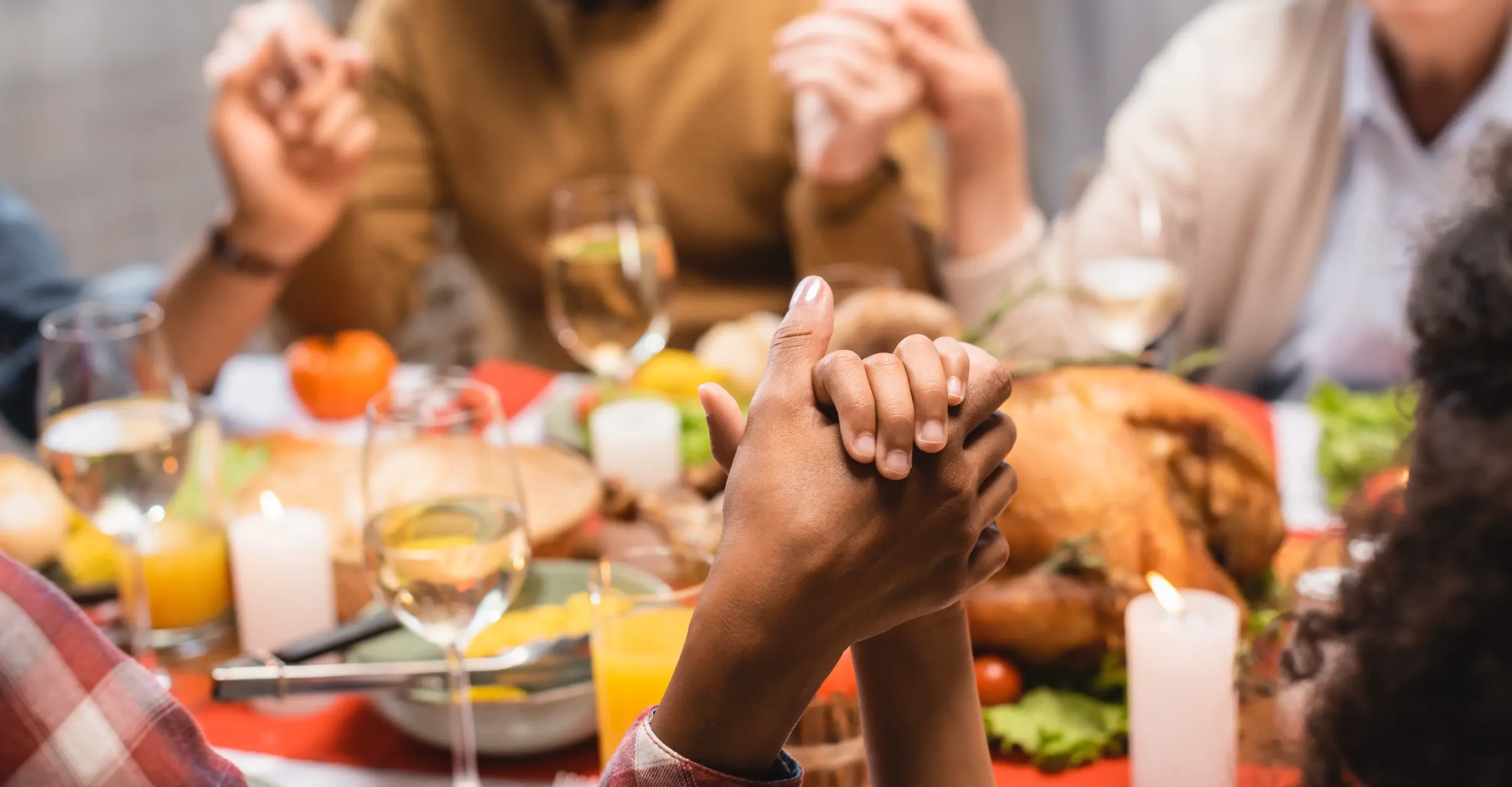 A family holding hands in prayer around a festive Thanksgiving dinner table. The focus is on two hands clasped in the foreground, while blurred figures of older adults and a roasted turkey are visible in the background. The table is adorned with candles, wine glasses, and autumn-themed dishes.