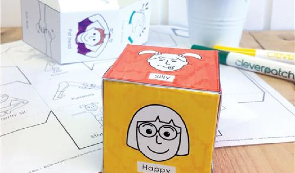 A paper cube with illustrated faces and emotion labels, such as "Silly" and "Happy," sits on a table with coloring supplies and a printout of similar cut-out templates.