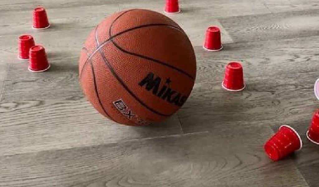 A basketball on a wooden floor surrounded by several small red plastic cups, some upright and some tipped over.
