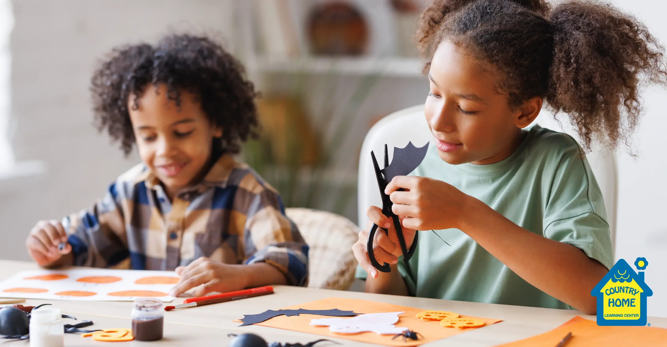 Two children making Halloween crafts with paper bats, pumpkins, and art supplies.