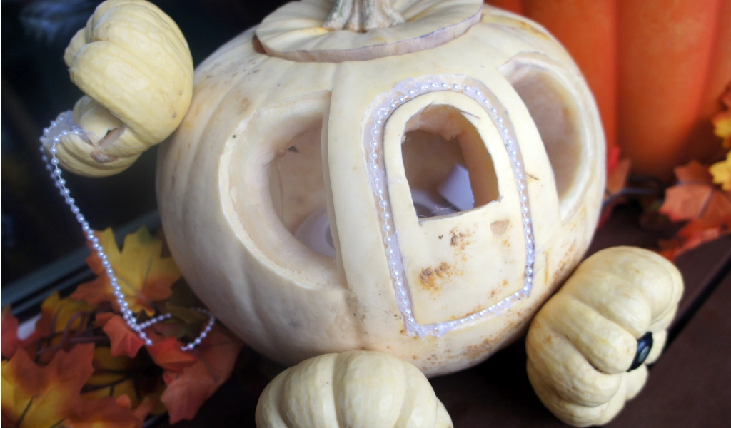 A white pumpkin carved and decorated to resemble a carriage, with small squash wheels and bead accents, surrounded by autumn leaves.