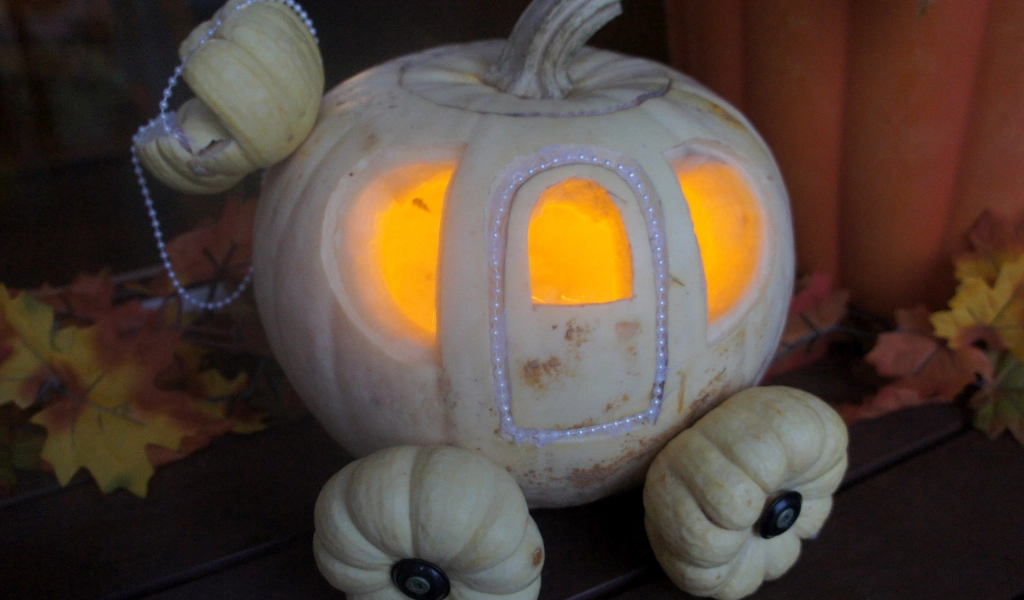 A white pumpkin carved and decorated to resemble a carriage, lit from inside, with small pumpkins as wheels and pearl-like beads outlining the windows.