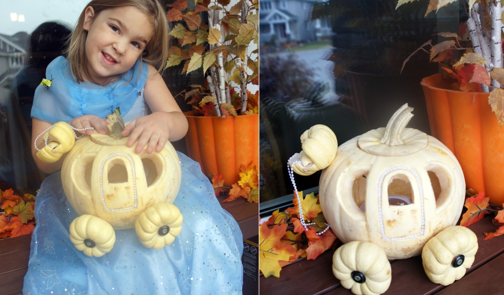 A young girl in a blue dress poses with a white pumpkin carved and decorated to resemble a carriage, surrounded by fall leaves and orange decorations.