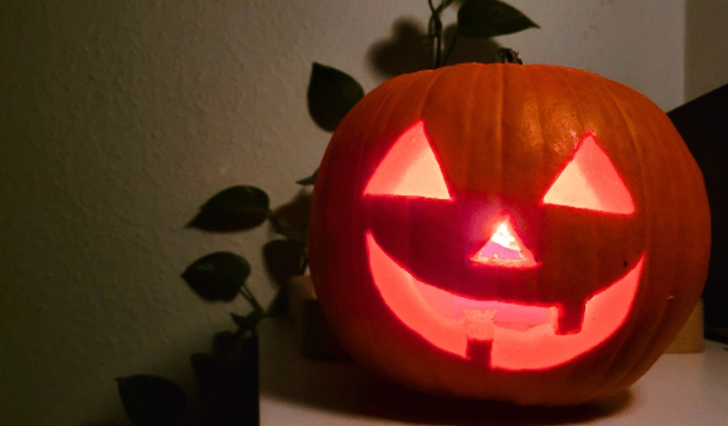 A carved pumpkin with a smiling face and triangle eyes is lit from inside, sitting on a white surface with plant leaves in the background.