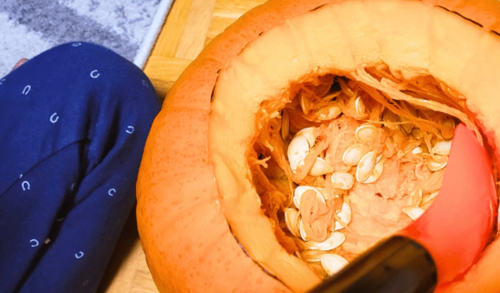 A pumpkin with its top cut off shows seeds and pulp inside, with a red scoop partially visible.