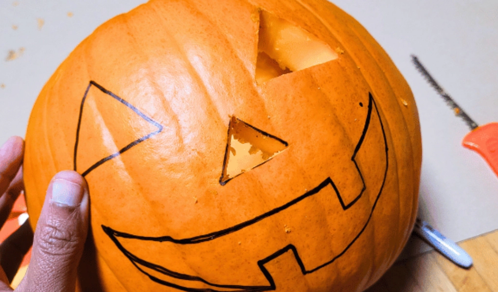 A pumpkin with a partially carved face and black marker outlines, held by a hand, sits next to an orange handled carving tool on a table.