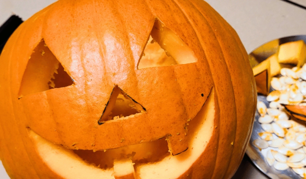 A carved pumpkin jack-o’-lantern with triangular eyes and a smiling mouth sits on a surface next to a plate with pumpkin seeds and pieces.