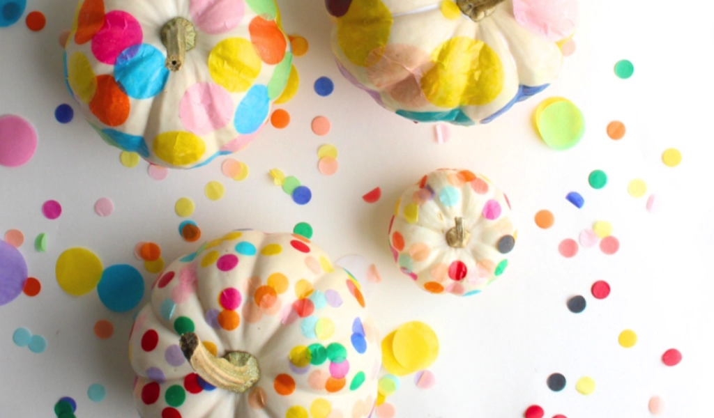 White pumpkins decorated with colorful confetti dots are arranged on a white surface scattered with more confetti.