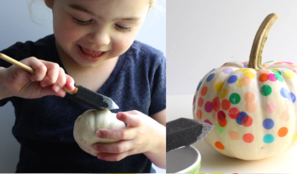 A child uses a foam brush to paint a small white pumpkin; next to them is a larger white pumpkin decorated with colorful confetti.