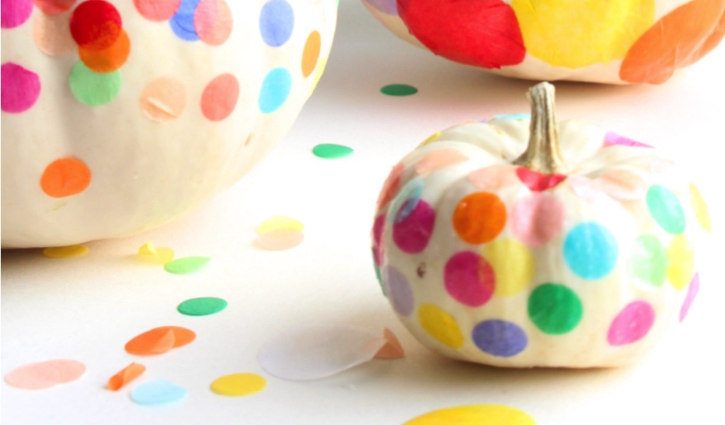 White pumpkins decorated with colorful confetti are placed on a white surface scattered with confetti.
