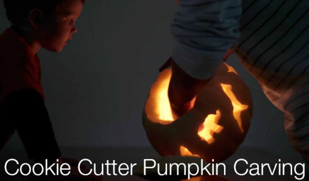 A child watches as an adult holds a lit pumpkin with cookie cutter-shaped carvings; the words "Cookie Cutter Pumpkin Carving" are overlaid at the bottom.