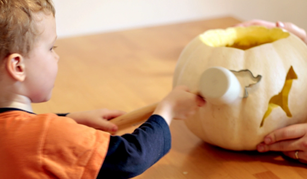A child uses a mallet to carve a design into a white pumpkin held by an adult on a wooden table.