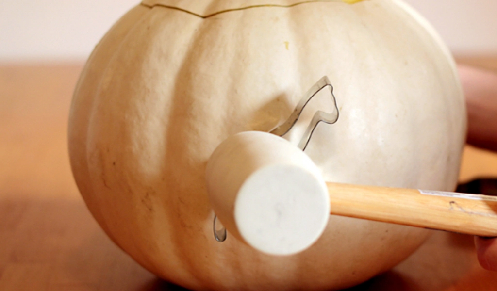 A person uses a mallet to tap a metal cookie cutter shaped like a horse into the side of a white pumpkin.