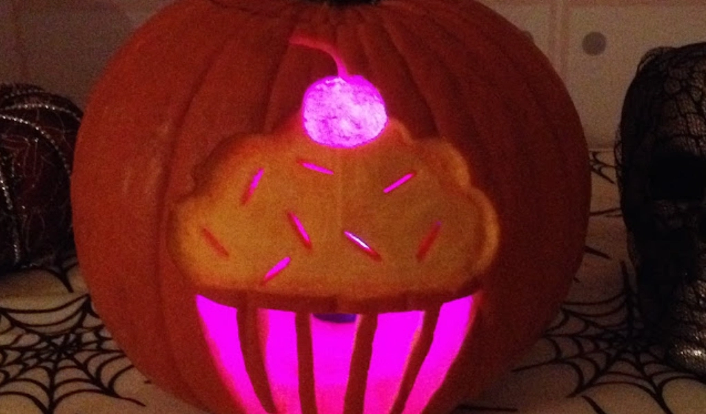 A pumpkin carved with a cupcake design and lit from inside with pink light, sitting on a table with spiderweb decorations.