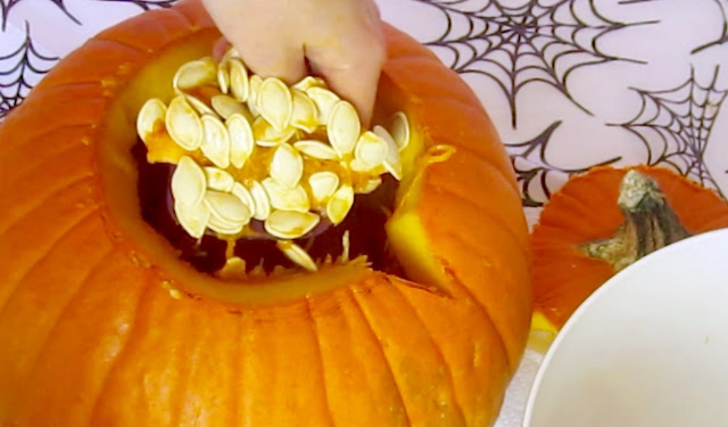 A hand scooping out pumpkin seeds and pulp from a carved pumpkin, with the lid removed and a bowl nearby. Spiderweb patterns are visible in the background.