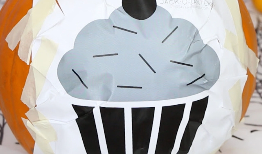 A black and white cupcake stencil is taped onto the surface of a pumpkin, likely as a guide for carving.