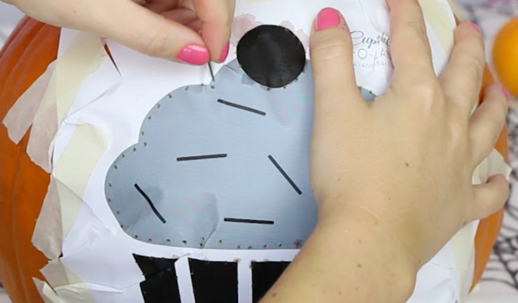 A person pins a cupcake stencil onto a pumpkin, preparing it for carving. The stencil is secured with tape and features dots along the outline.