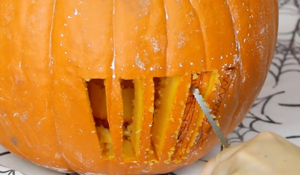 A hand is carving vertical rectangular openings into a pumpkin using a serrated knife, with dotted guide lines visible on the pumpkin's surface.