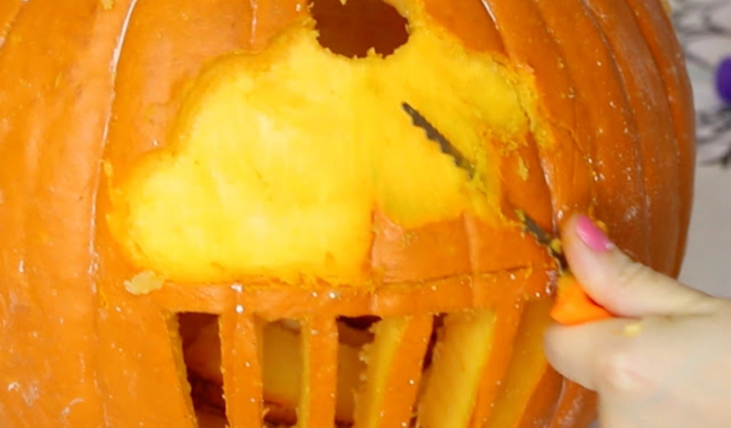 A hand uses a serrated tool to carve a pumpkin, cutting out a large section and vertical slots below it.