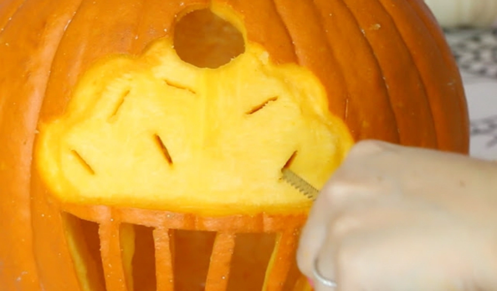 A hand carves a cupcake design into an orange pumpkin, with a round hole above the carved cupcake and vertical cuts below.