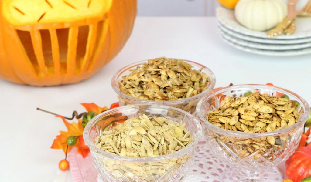 Three glass bowls filled with different roasted pumpkin seeds are displayed in front of a carved pumpkin and stacked white plates, with autumn leaves as decoration.