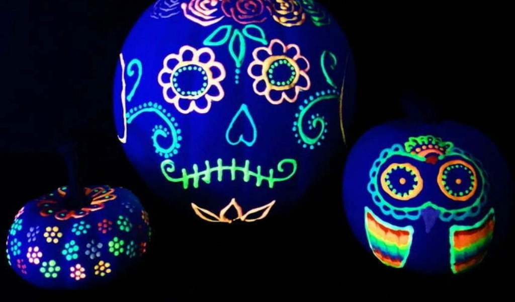 Three pumpkins decorated with neon, Day of the Dead-inspired designs glow under blacklight against a dark background.