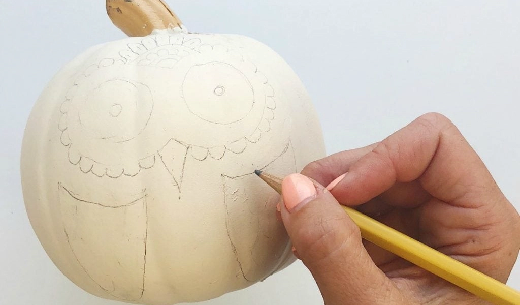 A hand uses a pencil to draw an owl face and wings on a white pumpkin.