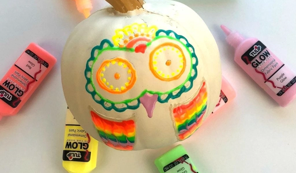 A white pumpkin decorated with neon puffy paint in the shape of an owl, surrounded by tubes of glow-in-the-dark paint.