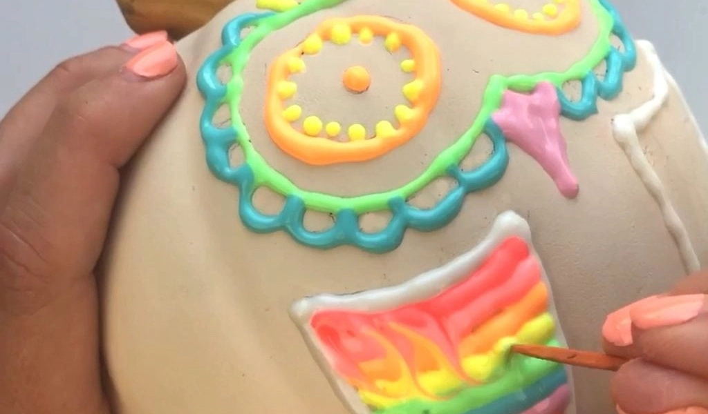 A person paints colorful designs with bright icing on a round, tan object, possibly a skull-shaped item, using a small brush.