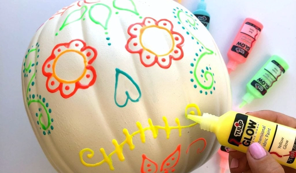 A white pumpkin is being decorated with neon glow paint in a sugar skull design; a hand applies yellow paint to create stitches on the mouth.