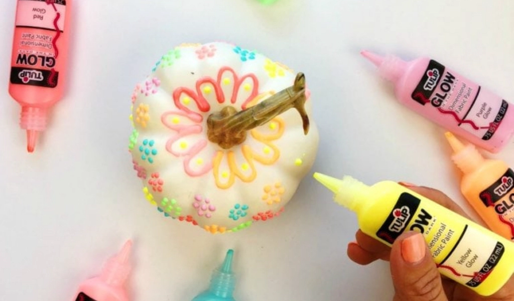 A hand decorates a white pumpkin with colorful glow-in-the-dark paints, surrounded by tubes of Tulip Glow paint in various colors.