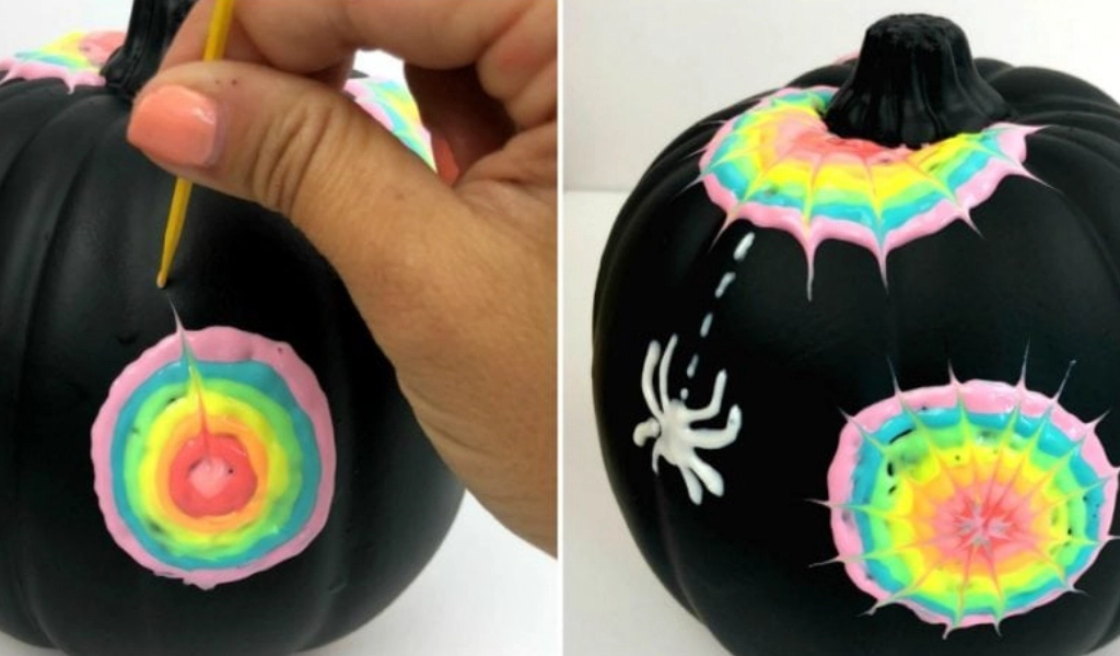 A hand uses a tool to drag paint on a black pumpkin, creating a rainbow web design, with a white plastic spider attached to the pumpkin.
