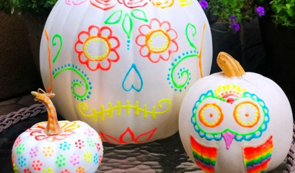 Three white pumpkins are decorated with colorful, Day of the Dead-inspired designs, featuring flowers, hearts, and an owl face, arranged on a table outdoors.