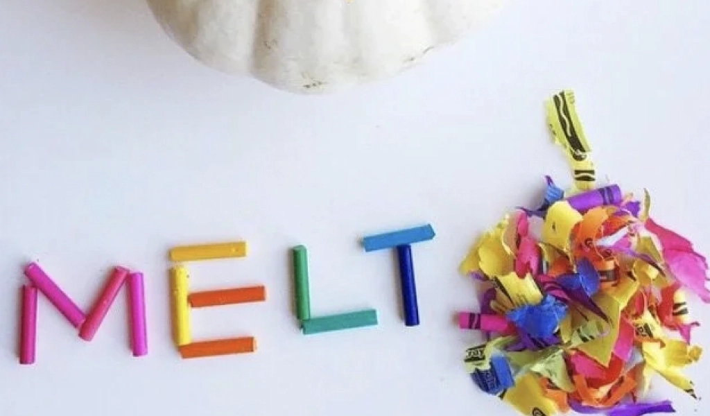 Broken crayon pieces spell the word "MELT" next to a pile of colorful crayon shavings on a white surface, with part of a white pumpkin visible at the top.