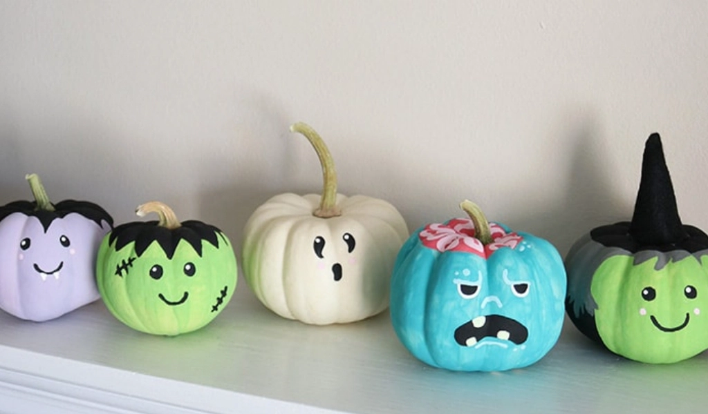 Five small pumpkins painted in pastel colors, each decorated with different cartoon faces and Halloween-themed designs, are lined up on a white surface against a neutral wall.