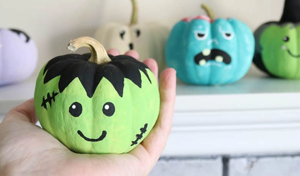 A hand holds a small green pumpkin painted with a smiling Frankenstein face; more painted pumpkins with various faces are on a shelf in the background.