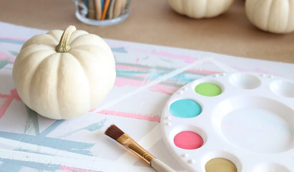 A white pumpkin, paintbrush, and paint palette with pastel colors on a table; more white pumpkins and a jar of brushes are in the background.