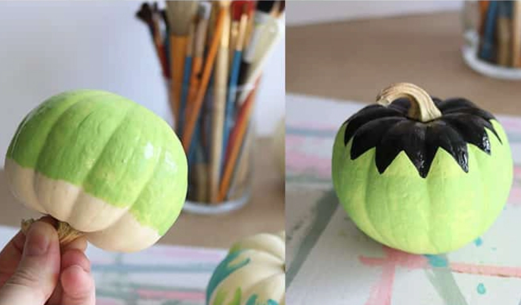 A small pumpkin is painted light green on one side and green with a black zigzag pattern near the stem on the other.