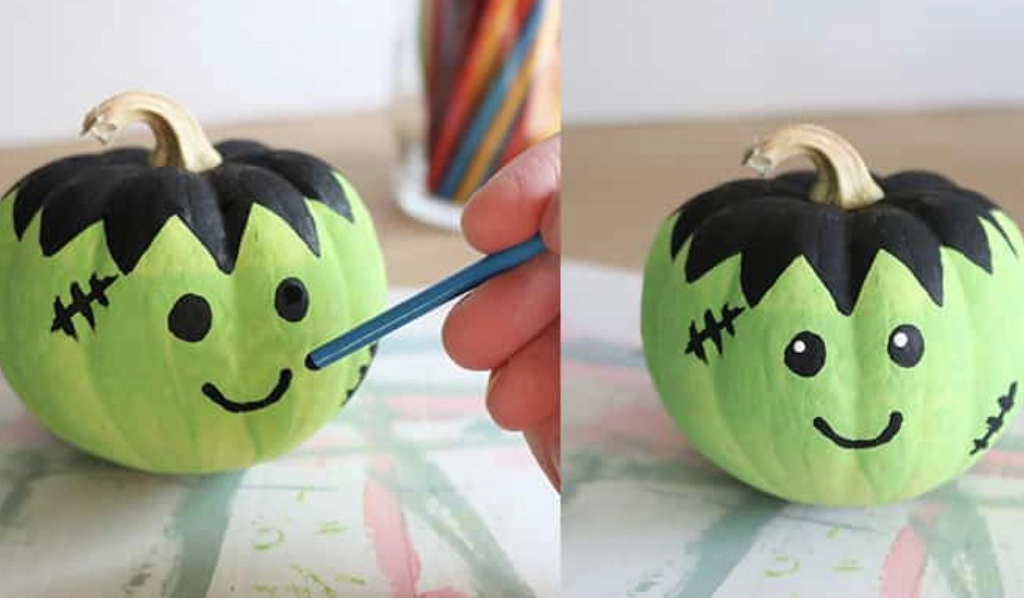 Two small pumpkins painted light green with black “hair,” stitches, and smiling faces; a hand uses a brush to paint details on one of the pumpkins.