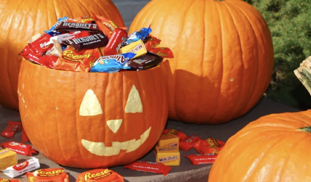 A carved pumpkin filled with assorted candy bars sits on a table surrounded by other uncarved pumpkins and scattered candy.