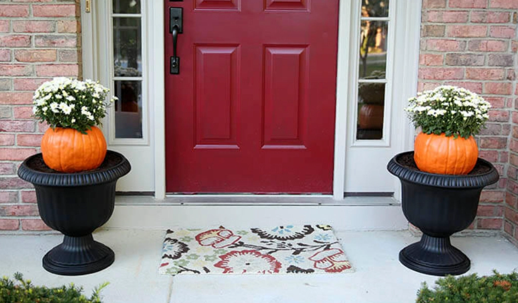Two black planters with orange pumpkins and white flowers sit on either side of a red front door, with a patterned doormat on the porch.